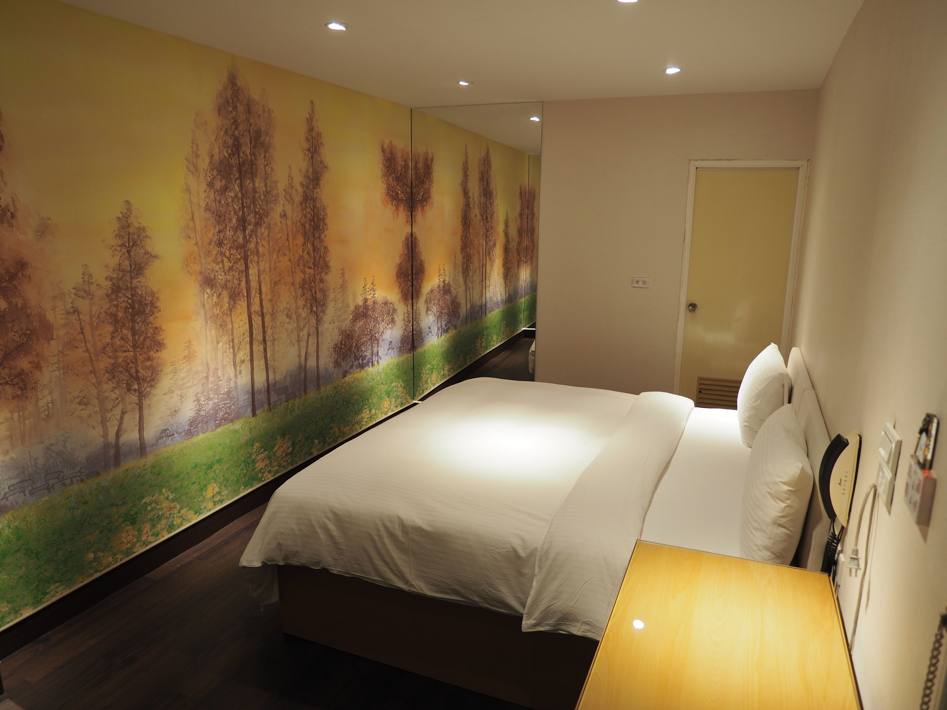 A bedroom with a large bed, a nightstand, a wall-mounted lamp, and a door.