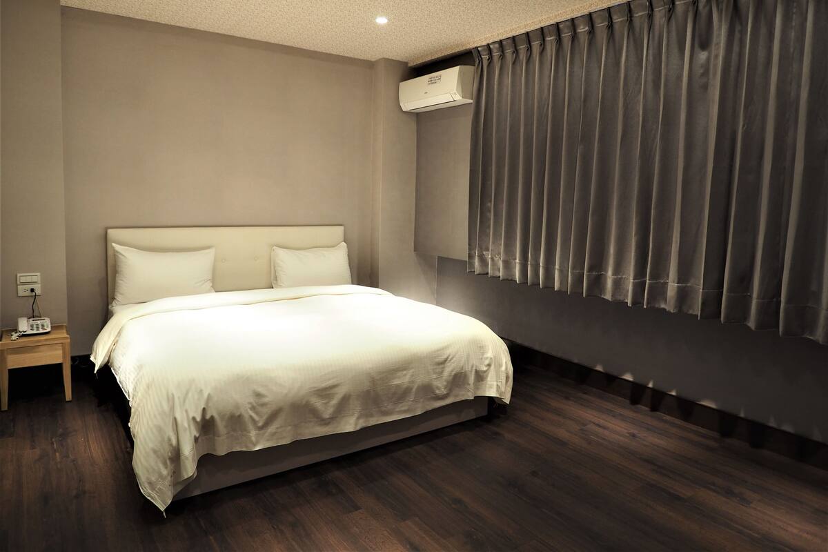 Un dormitorio con una cama grande, una mesita de noche, un equipo de aire acondicionado y una ventana con cortinas.