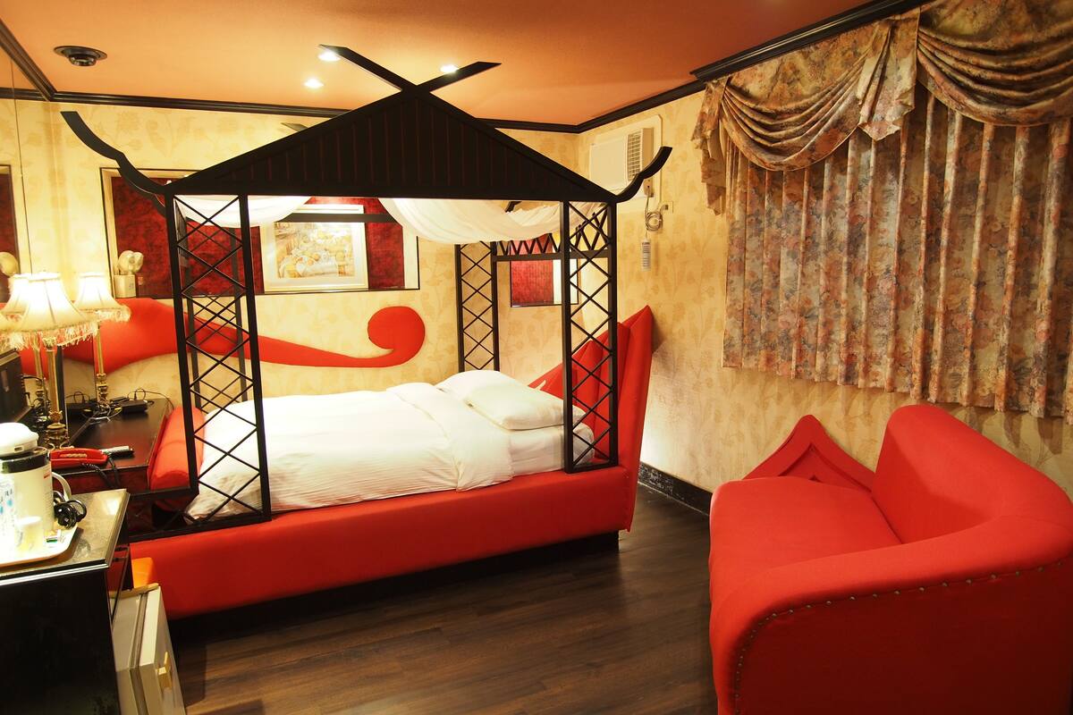 Una habitación con cama con dosel, un sillón rojo, un escritorio con lámpara y un teléfono.