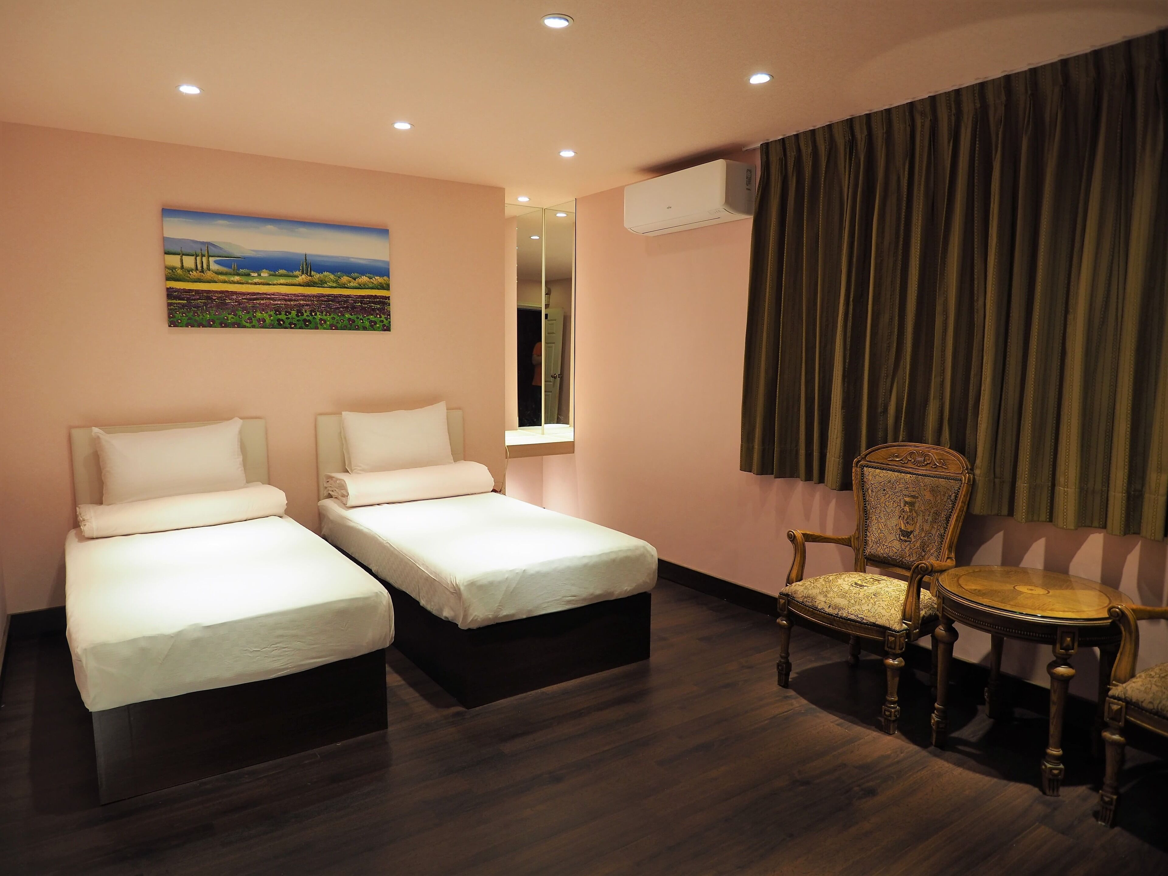 A hotel room with two single beds, a wooden chair, a small round table, a painting on the wall, and an air conditioning unit.