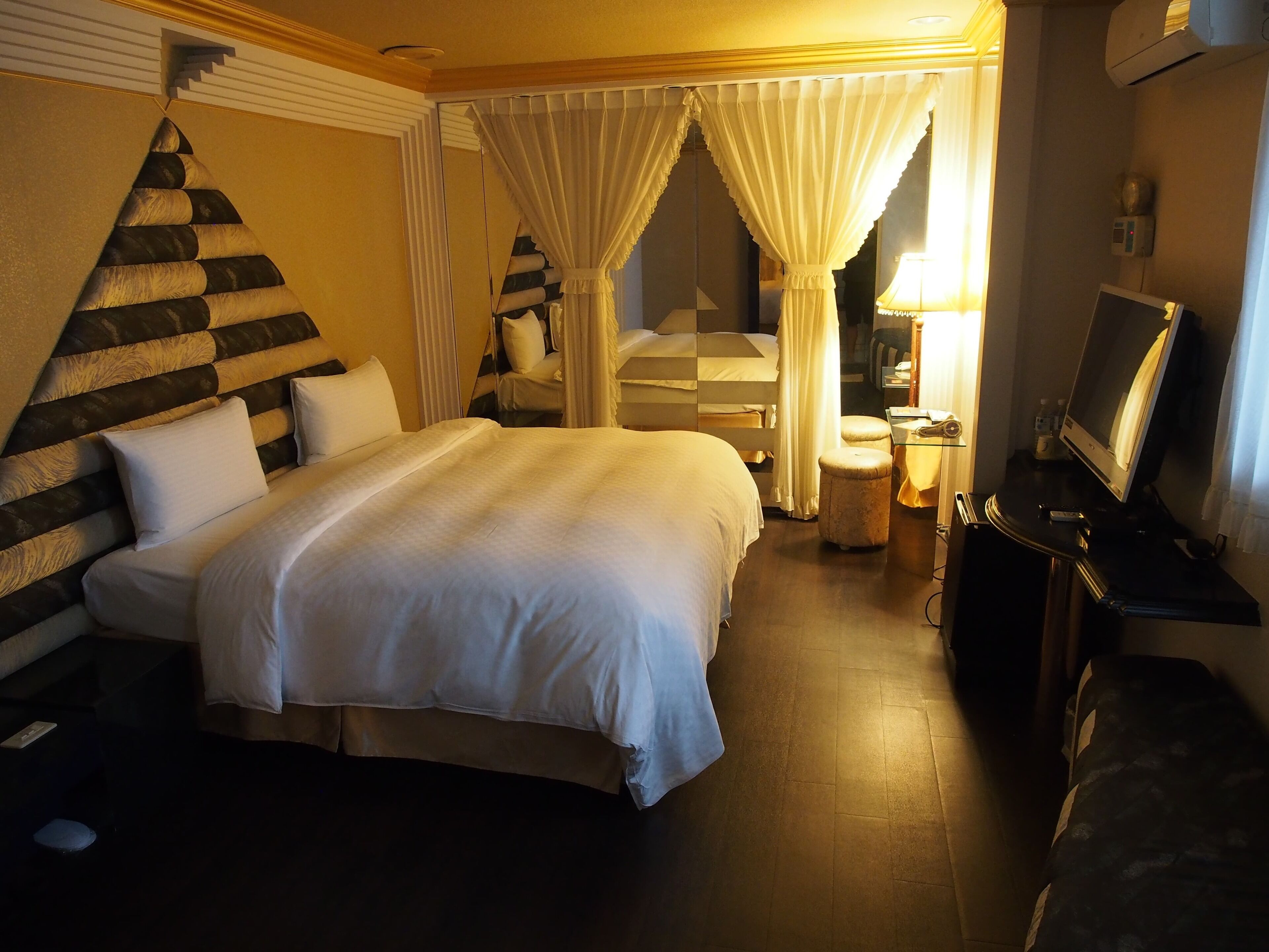 A hotel room with a large bed, a desk with a lamp, a television, and a mirror.