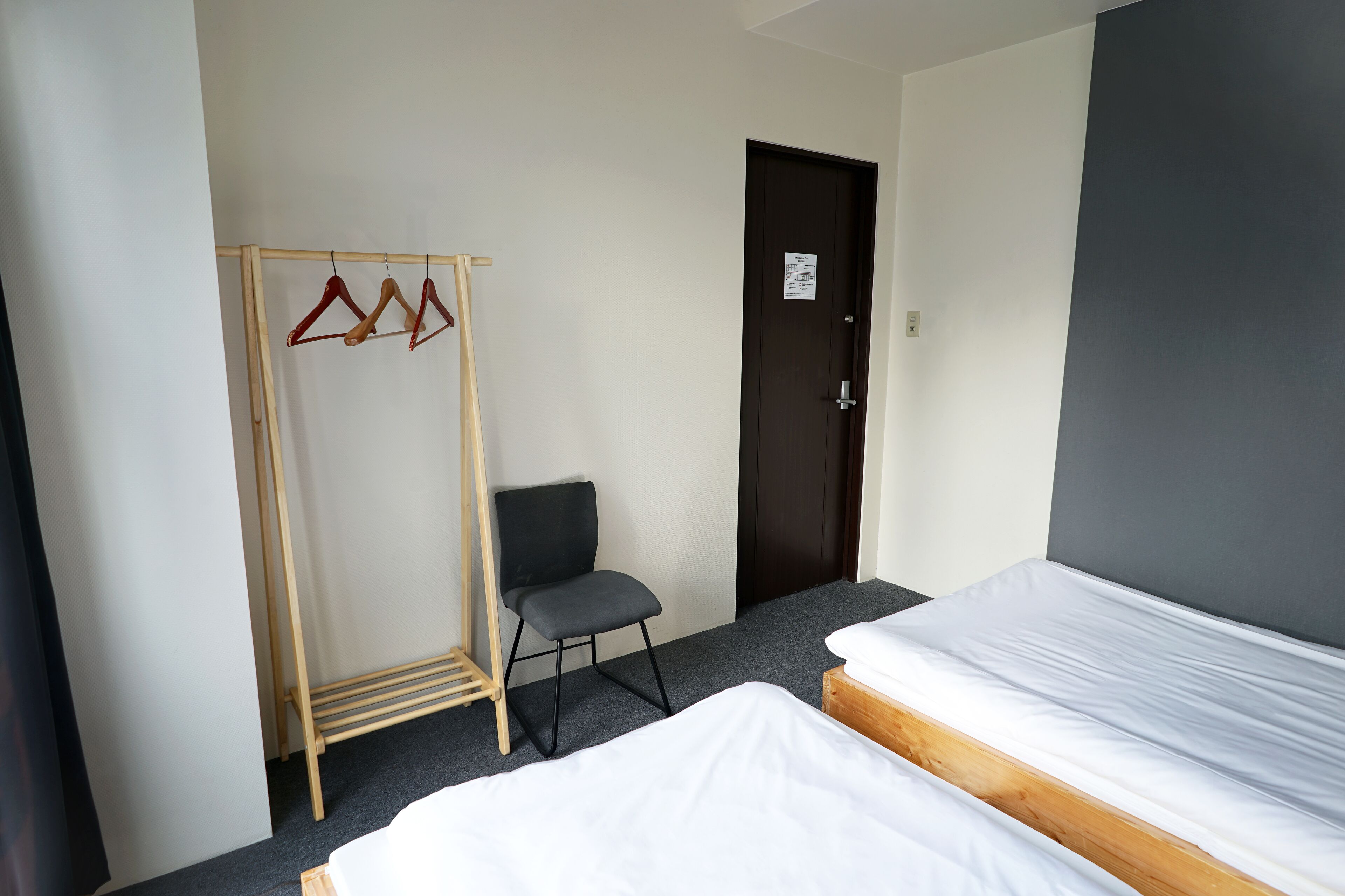 A room with two beds, a wooden drying rack with clothes, a chair, and a door.