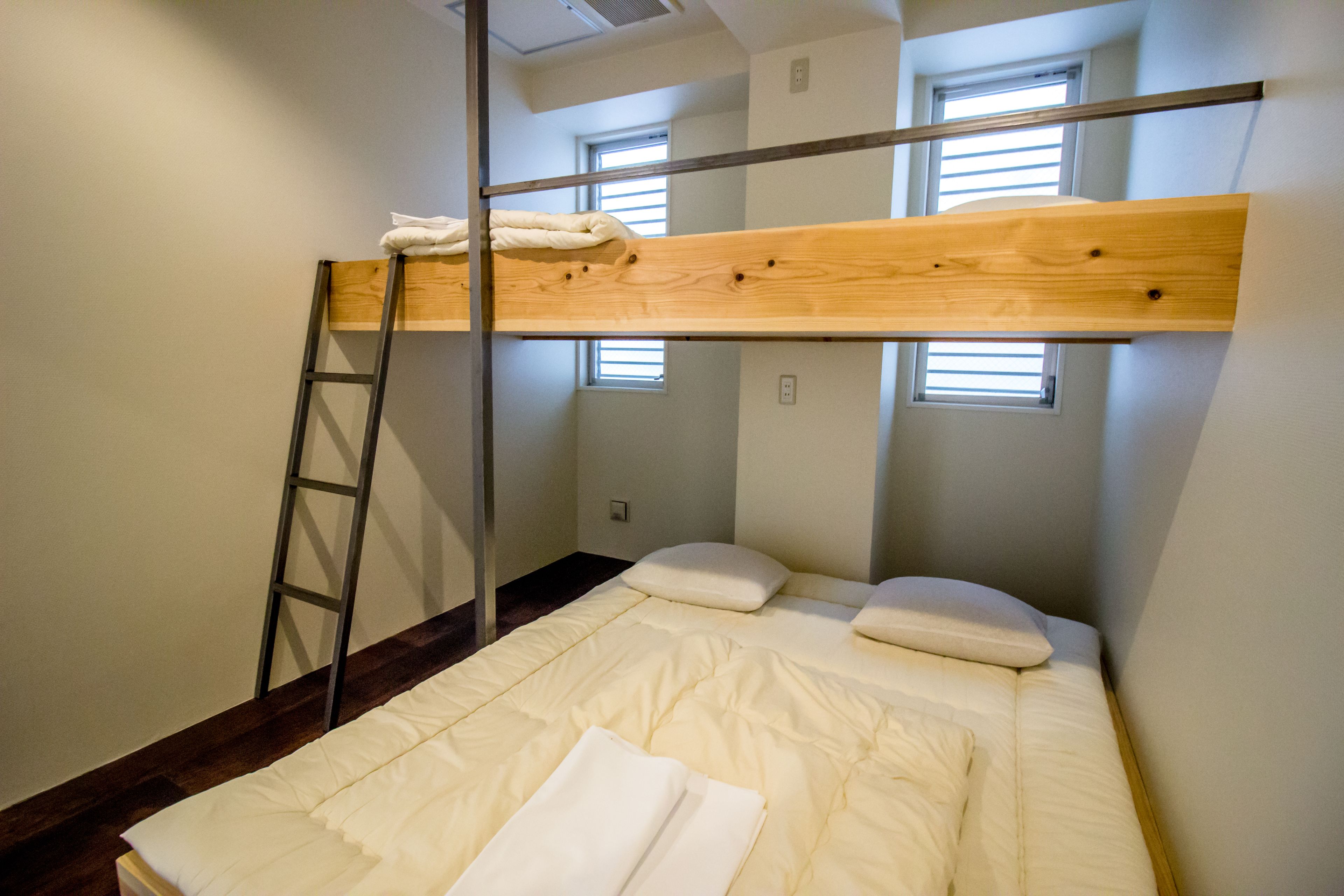A bunk bed with a wooden frame and white bedding, a ladder, and two windows with blinds.