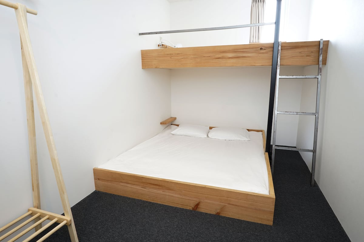 Una habitación pequeña con una cama individual y una litera, una escalera metálica y un tendedero de madera.