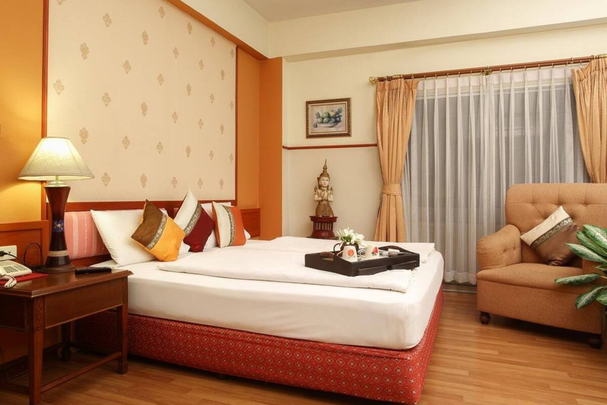 A hotel room with a bed, a chair, a nightstand, a lamp, a statue, a picture on the wall, and a window with curtains.