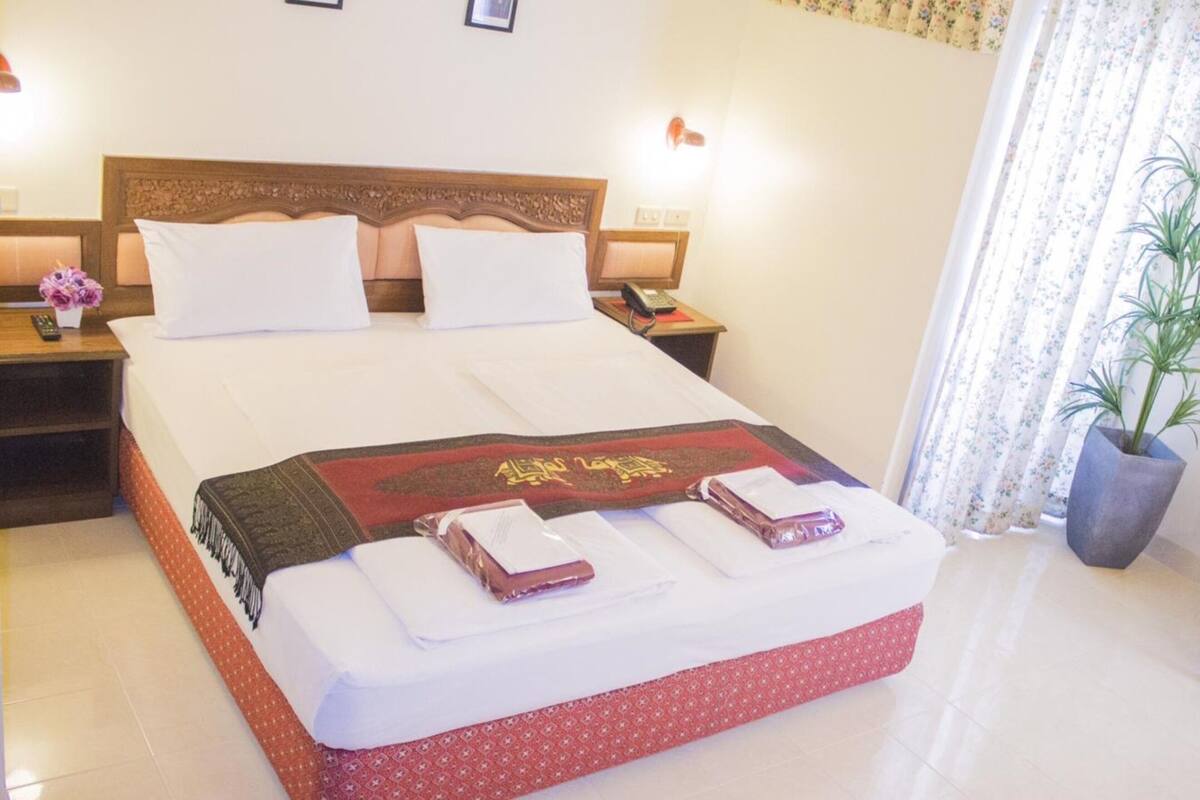 A neatly made bed with a red and gold decorative throw, white pillows, and neatly folded towels. There are two bedside tables with lamps and a phone. The room has a floral curtain and a potted plant.