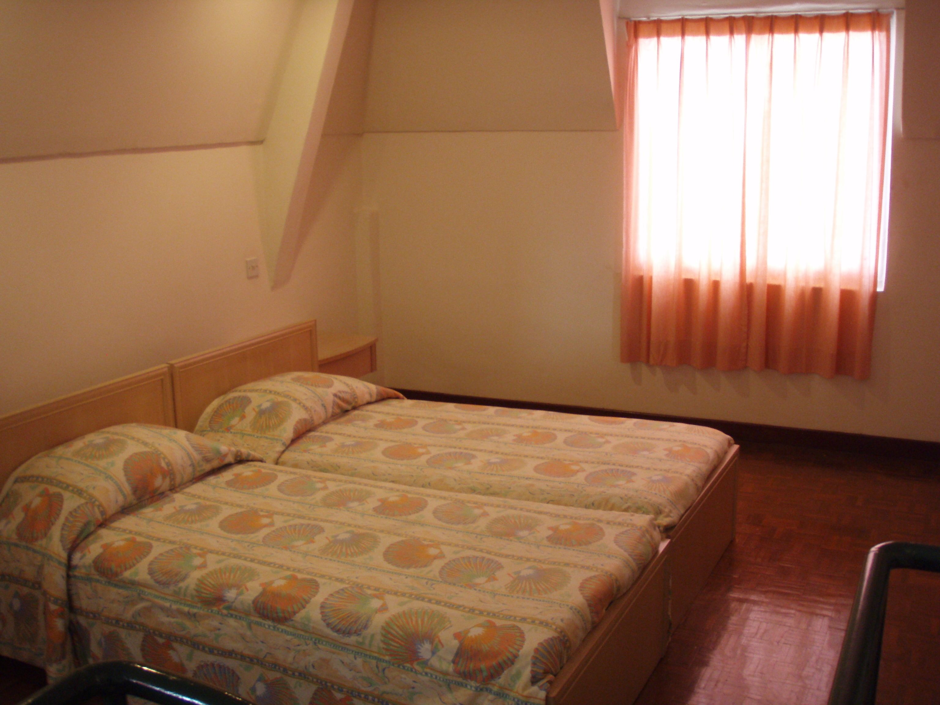 Ein kleines, schlichtes Zimmer mit einem Bett, einem Stuhl und einem Fenster mit Vorhängen.