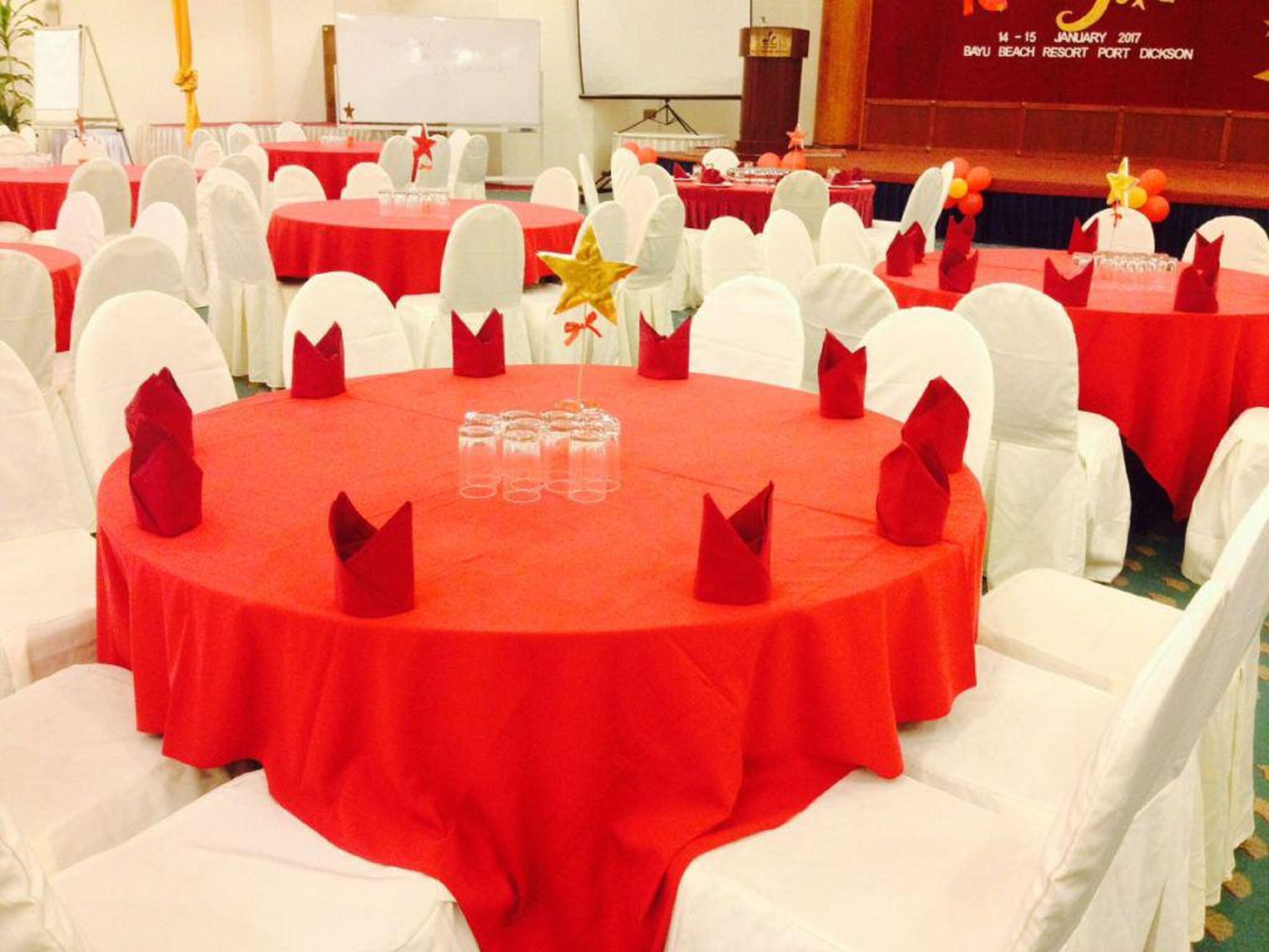 Salão de banquetes com mesas redondas cobertas por toalhas vermelhas, cadeiras brancas e guardanapos vermelhos.