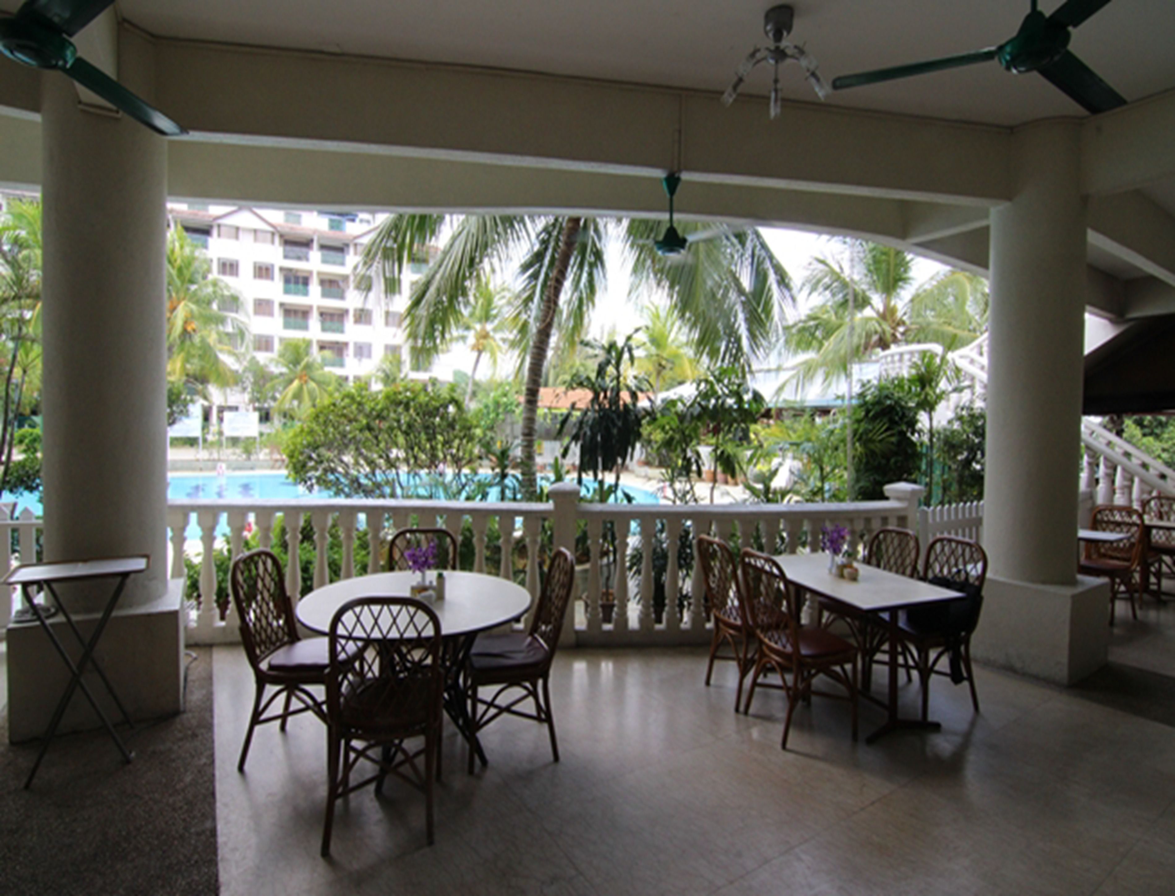 A covered outdoor area with tables and chairs, a pool in the background, and palm trees.