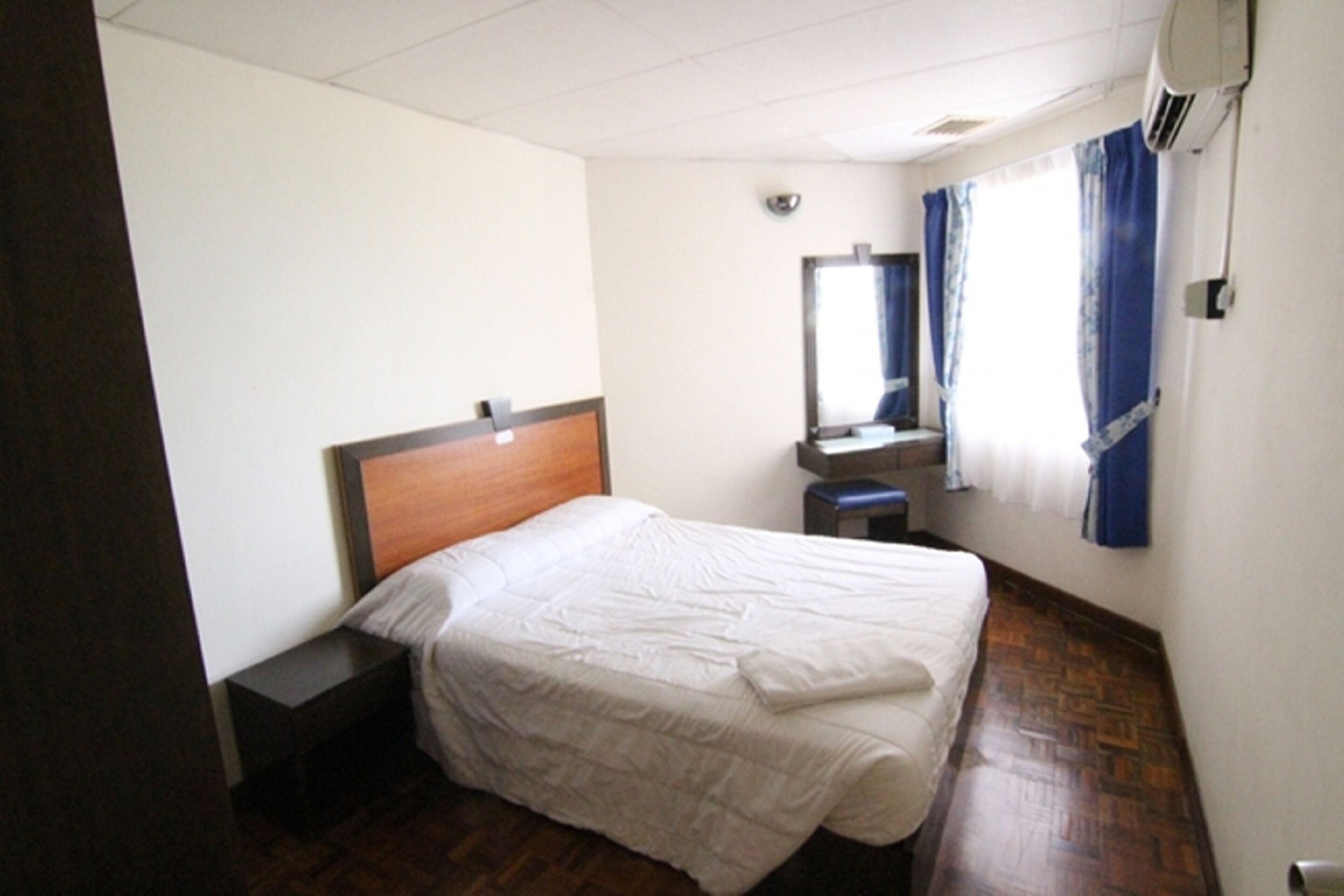 A hotel room with a wooden headboard bed, a small bedside table, a mirror, and a window with curtains.