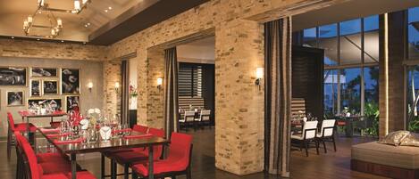 A spacious dining area with red chairs and a long table, brick walls, and large windows offering a view of the outdoors.