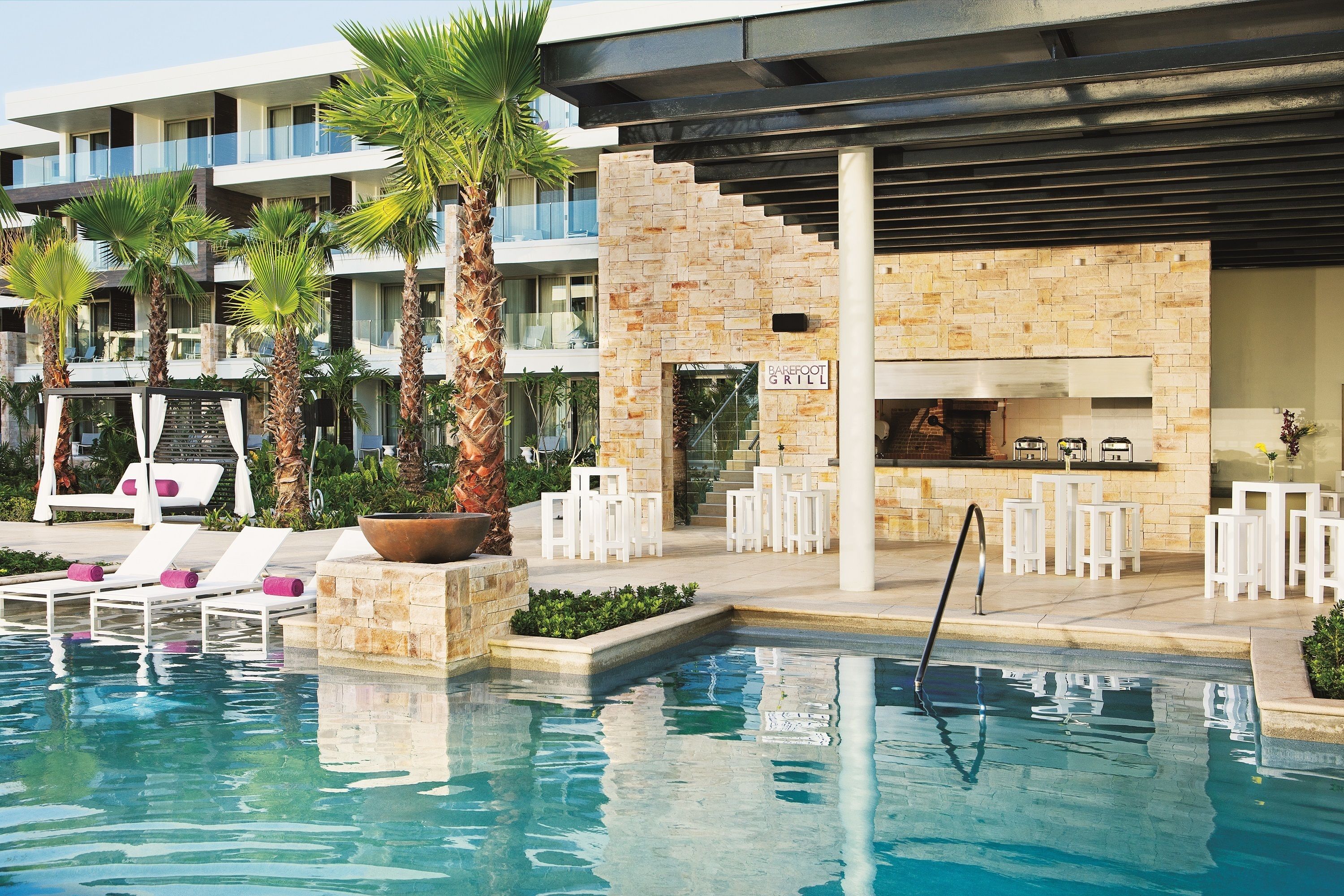 A pool area with lounge chairs, palm trees, and a bar area with outdoor seating.