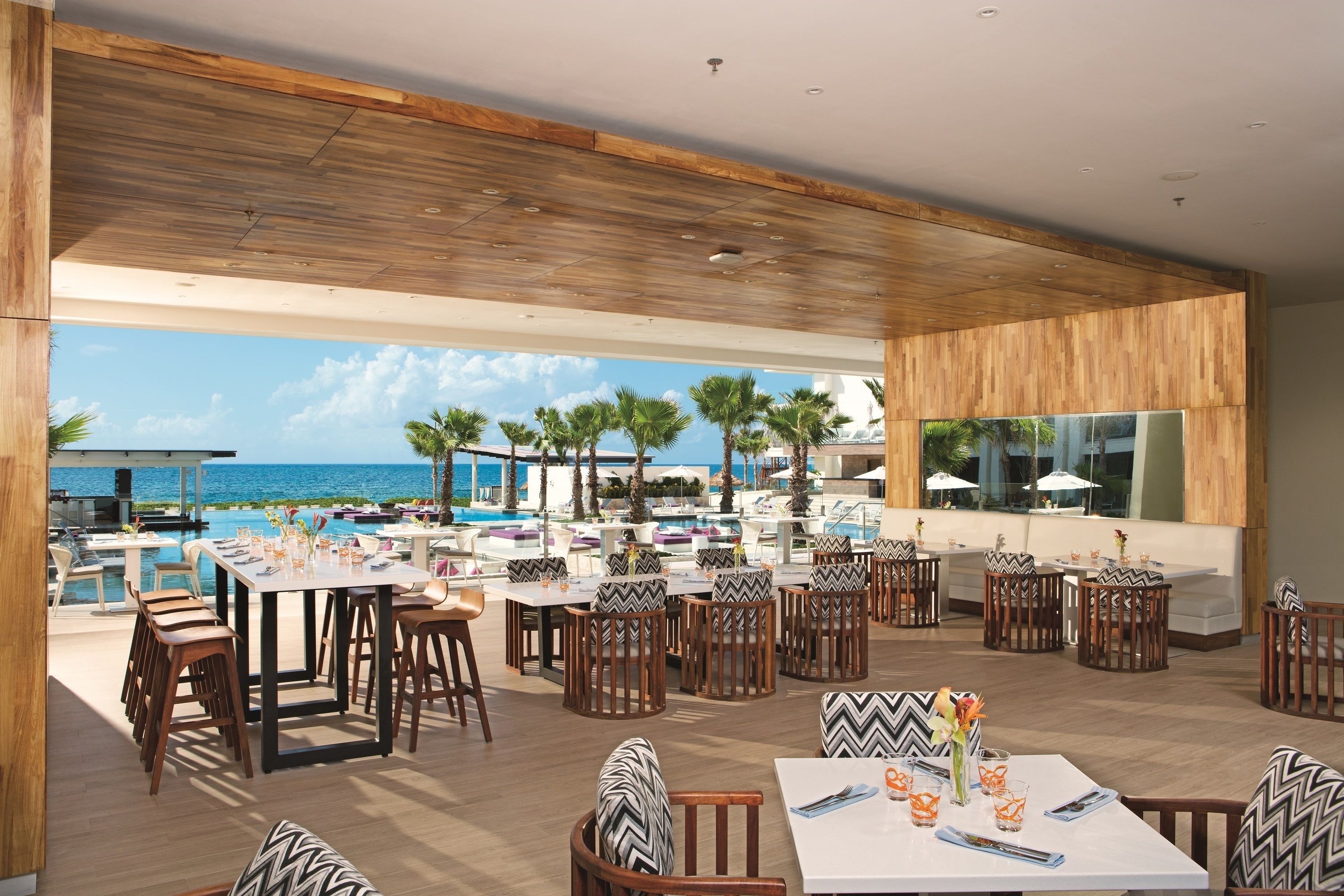 An outdoor dining area with wooden tables and chairs, overlooking a pool and the ocean.