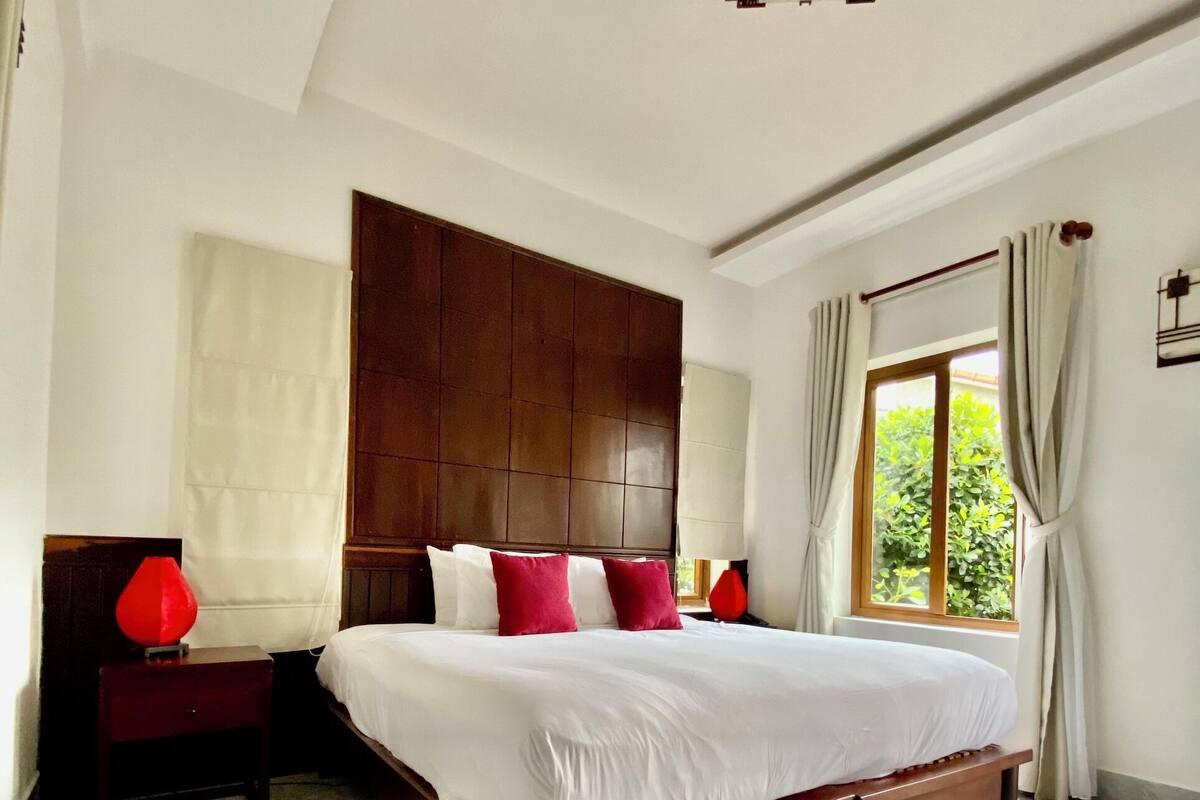 A bedroom with a wooden bed, white bedding, red pillows, a nightstand, a lamp, a window with curtains, and a view of greenery outside.