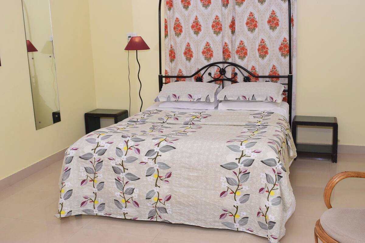 A bedroom with a bed, two bedside tables, a chair, a mirror, and a window with floral curtains.