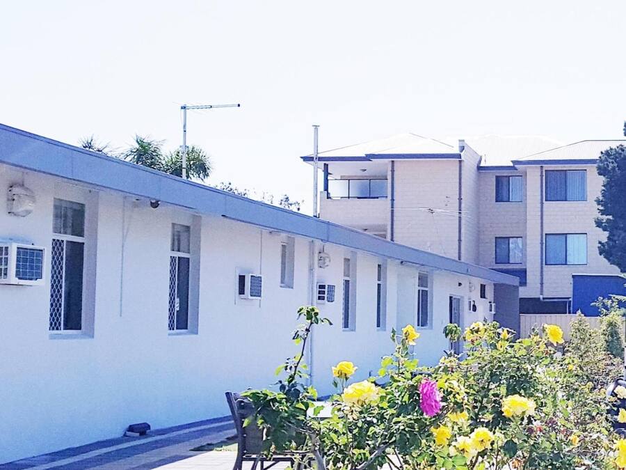 A row of white buildings with multiple windows, a paved walkway, and some greenery in the foreground.