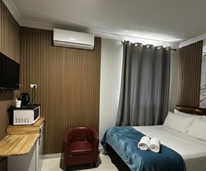A hotel room with a bed, a desk with a microwave, a chair, an air conditioner, and a curtain. - Perth City Motel (Thornlie)