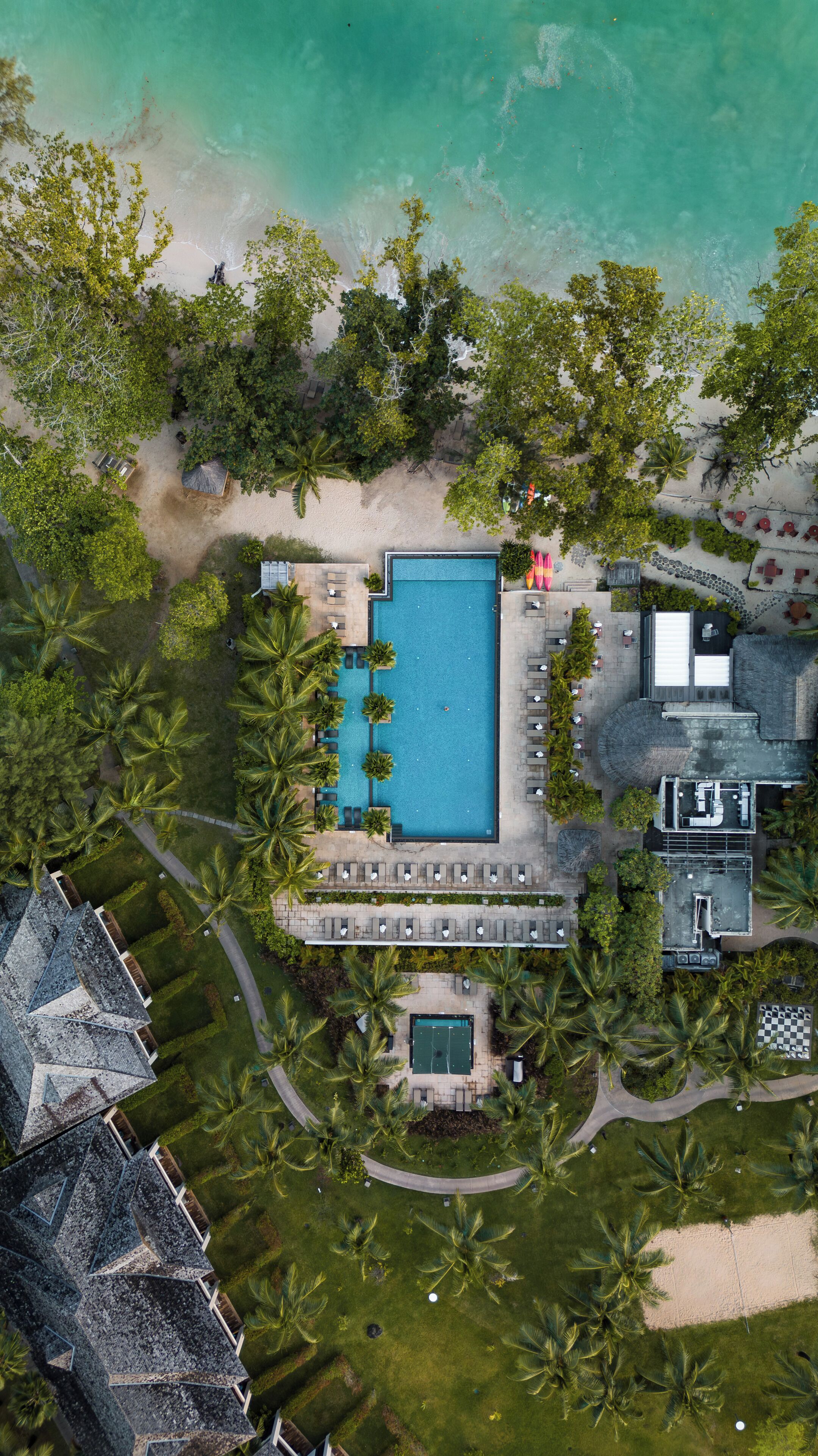 Vista aérea de un resort con una piscina central, rodeado de palmeras y una playa.
