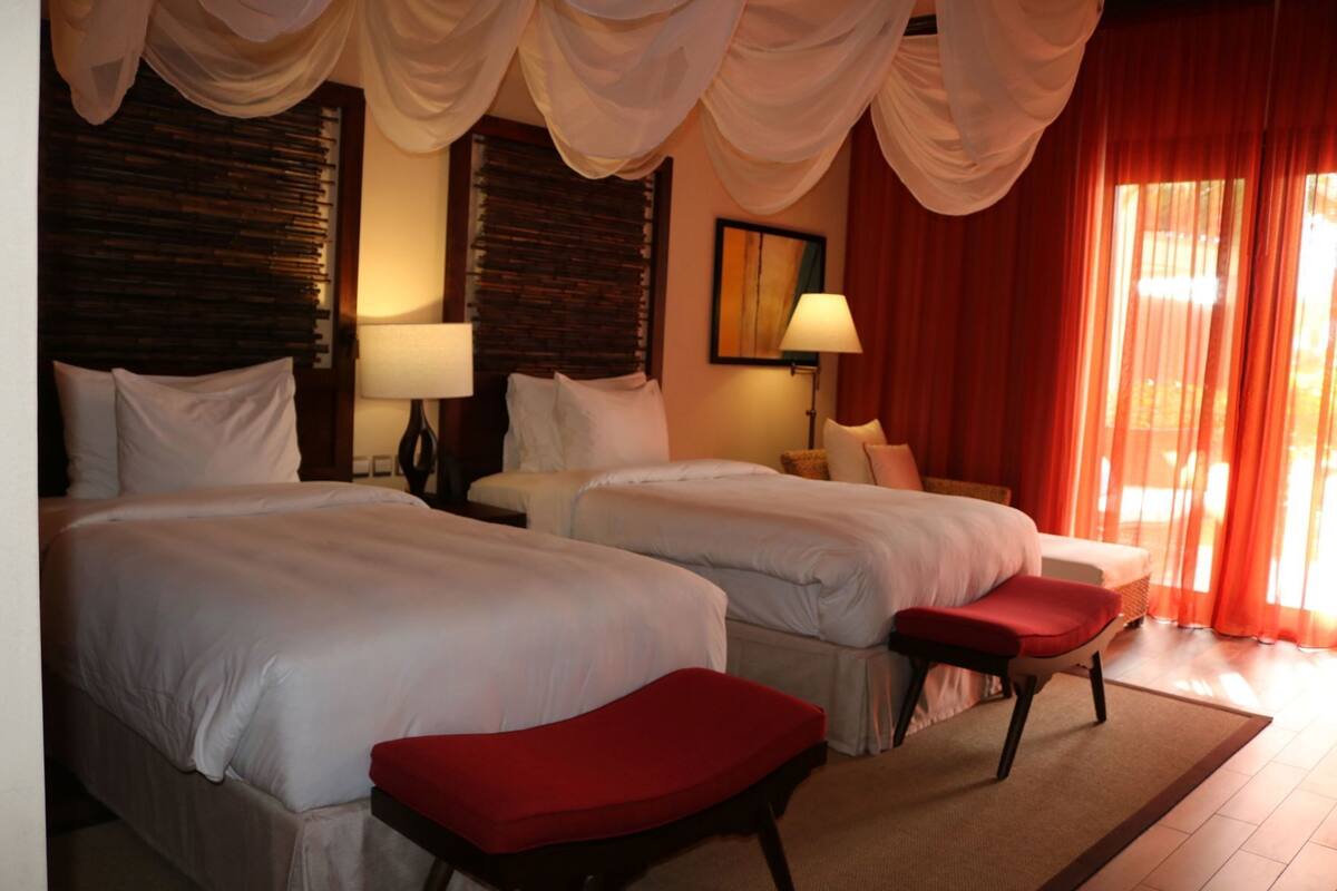 A hotel room with two beds, a red bench, and a view of the outdoors.