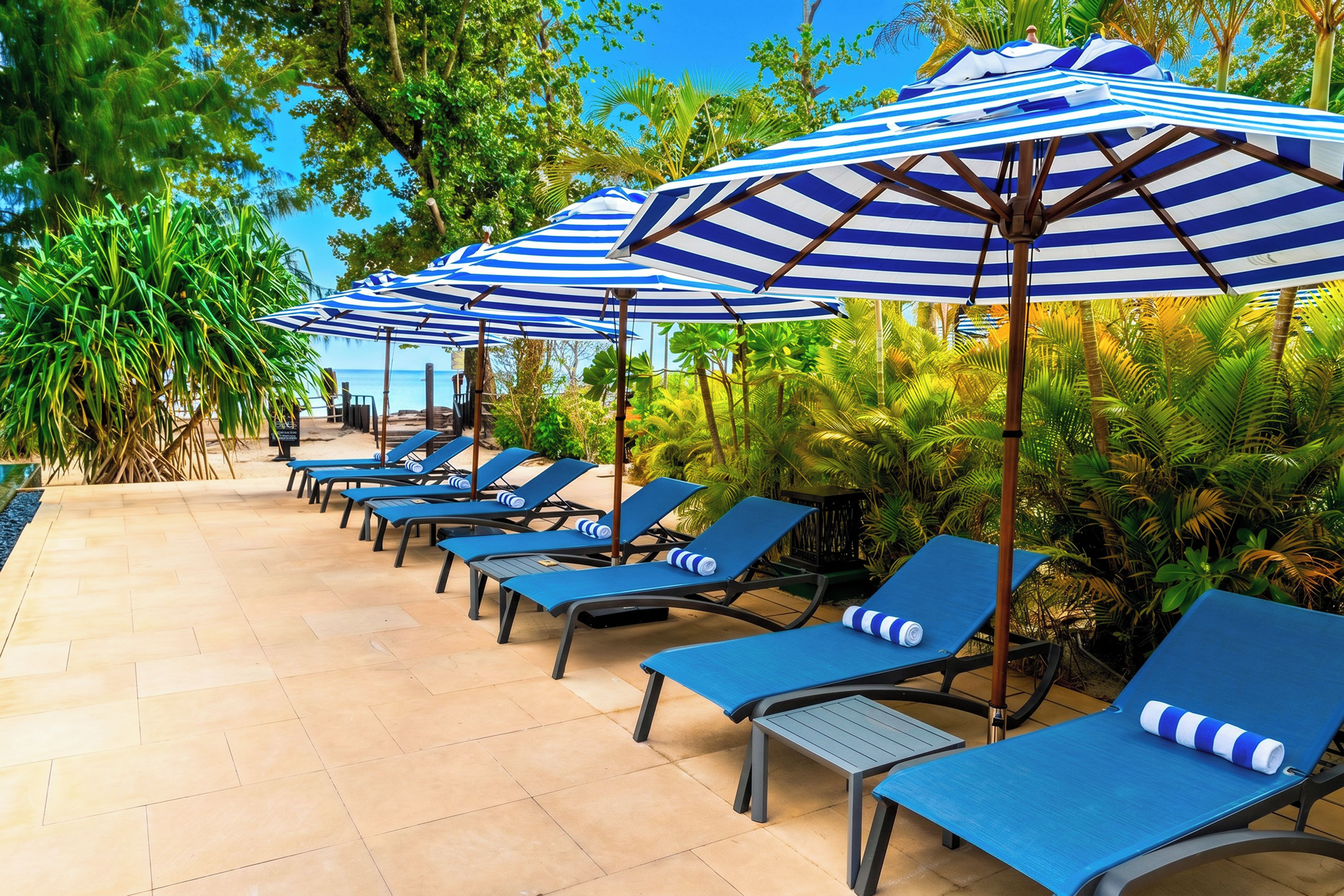 Une rangée de fauteuils de jardin bleus, surmontés de parasols à rayures, est disposée sur une terrasse carrelée, avec des palmiers en arrière-plan.