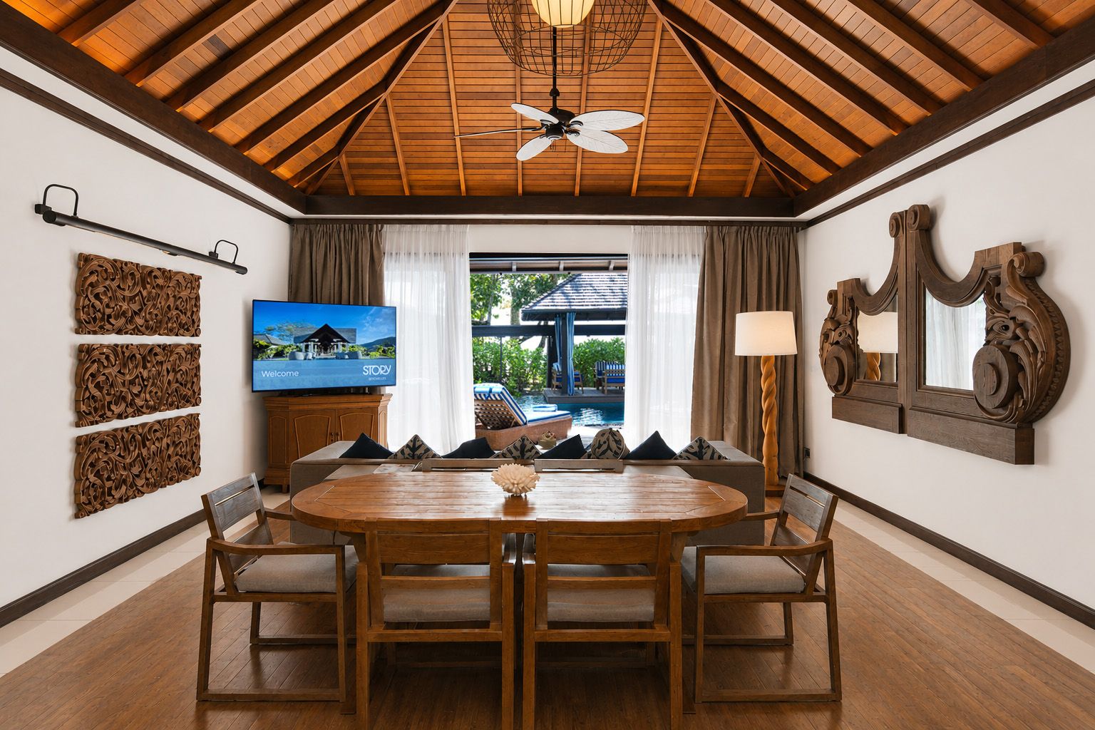 A dining area with a wooden table and chairs, a television on the wall, and a view of an outdoor pool.