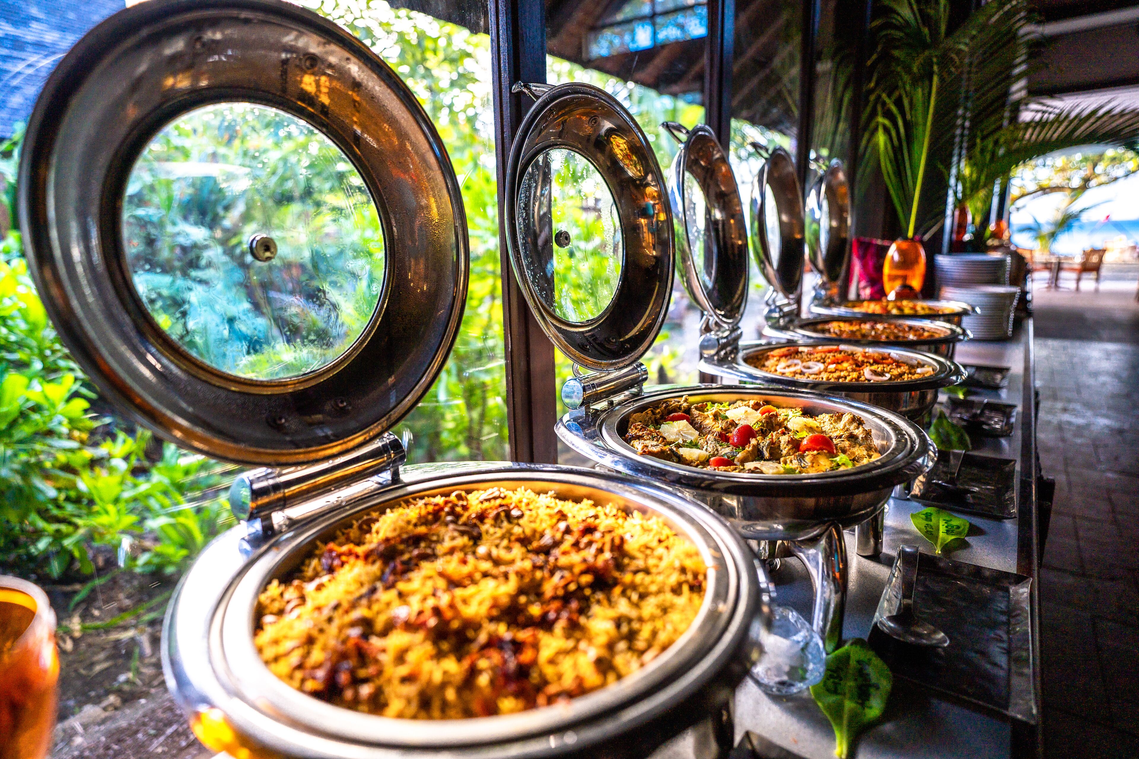 Ein Buffet mit verschiedenen Speisen, darunter Reis und Fleisch, draußen mit Blick auf Grünflächen und ein Gebäude im Hintergrund.