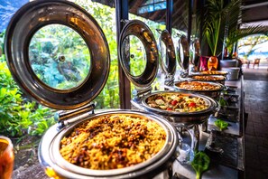 A buffet with various dishes, including rice and meat, set up outdoors with a view of greenery and a building in the background.