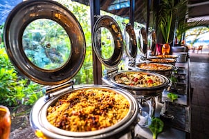 A buffet with various dishes, including rice and meat, set up outdoors with a view of greenery and a building in the background.