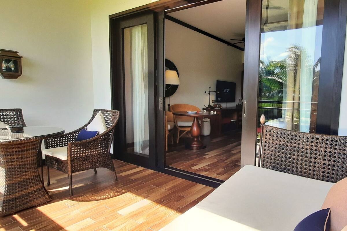 A room with a glass door leading to a balcony, a wicker sofa, a small table, and a TV in the background.