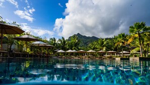 A clear blue pool with lounge chairs and umbrellas, surrounded by palm trees and a mountain in the background.