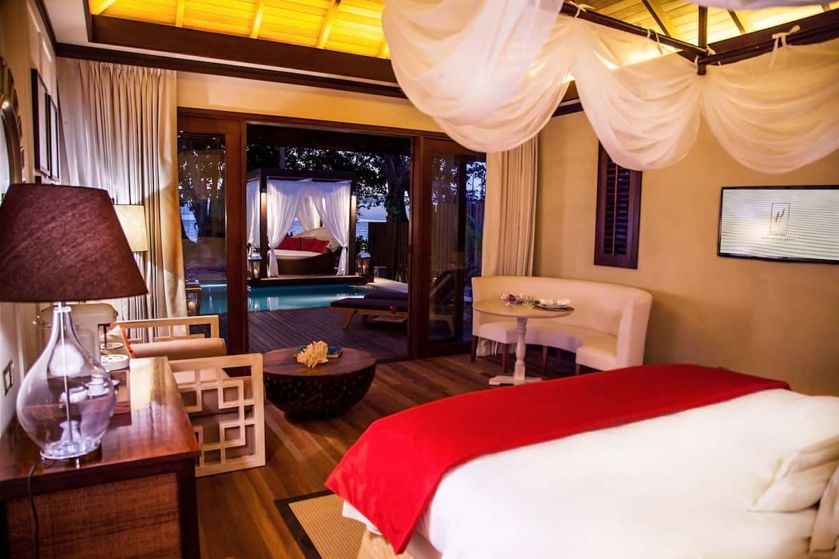 A hotel room with a bed, a desk with a lamp, a small table, a sofa, and a view of an outdoor swimming pool.