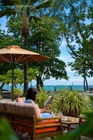 A person relaxing on a striped sofa under a large umbrella, with a view of the sea and lush greenery.