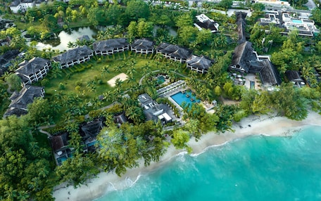 An aerial view of a resort with a circular layout of villas, a central pool, and a beach.