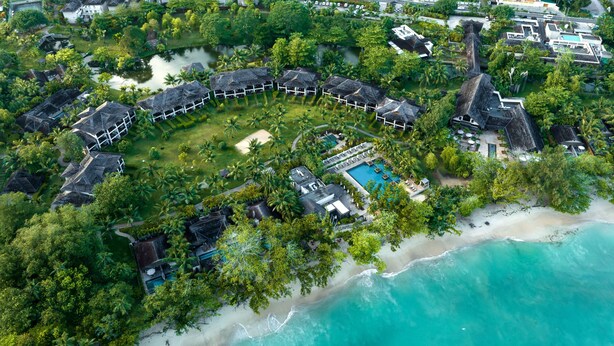 An aerial view of a resort with a circular layout of villas, a central pool, and a beach.