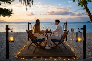 Una pareja cenando en una mesa de madera en una terraza de playa, con faroles y velas, con vista al mar.