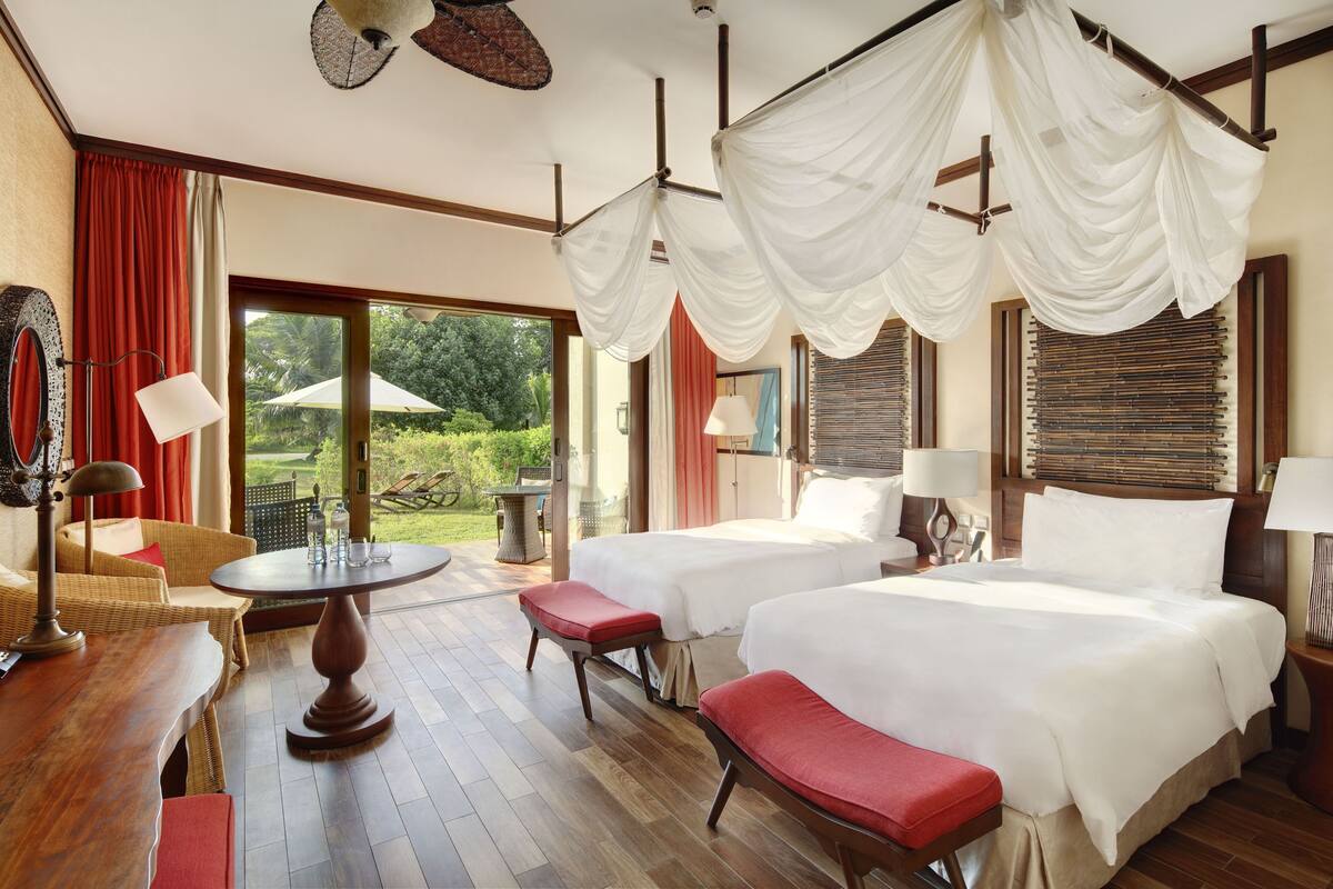 A hotel room with two beds, a wooden table, a wicker chair, and a balcony with a view of greenery.