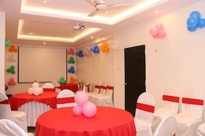 Une salle de conférence avec des tables rondes, des chaises blanches ornées de rubans rouges et des ballons colorés accrochés aux murs.