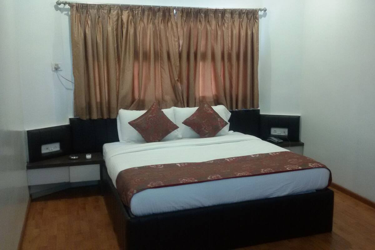 Uma cama bem arrumada com colcha estampada, duas mesas de cabeceira com abajures e uma janela com cortinas.