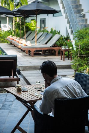 Un hombre sentado en una mesa al aire libre con vista a una piscina y sillones de descanso.