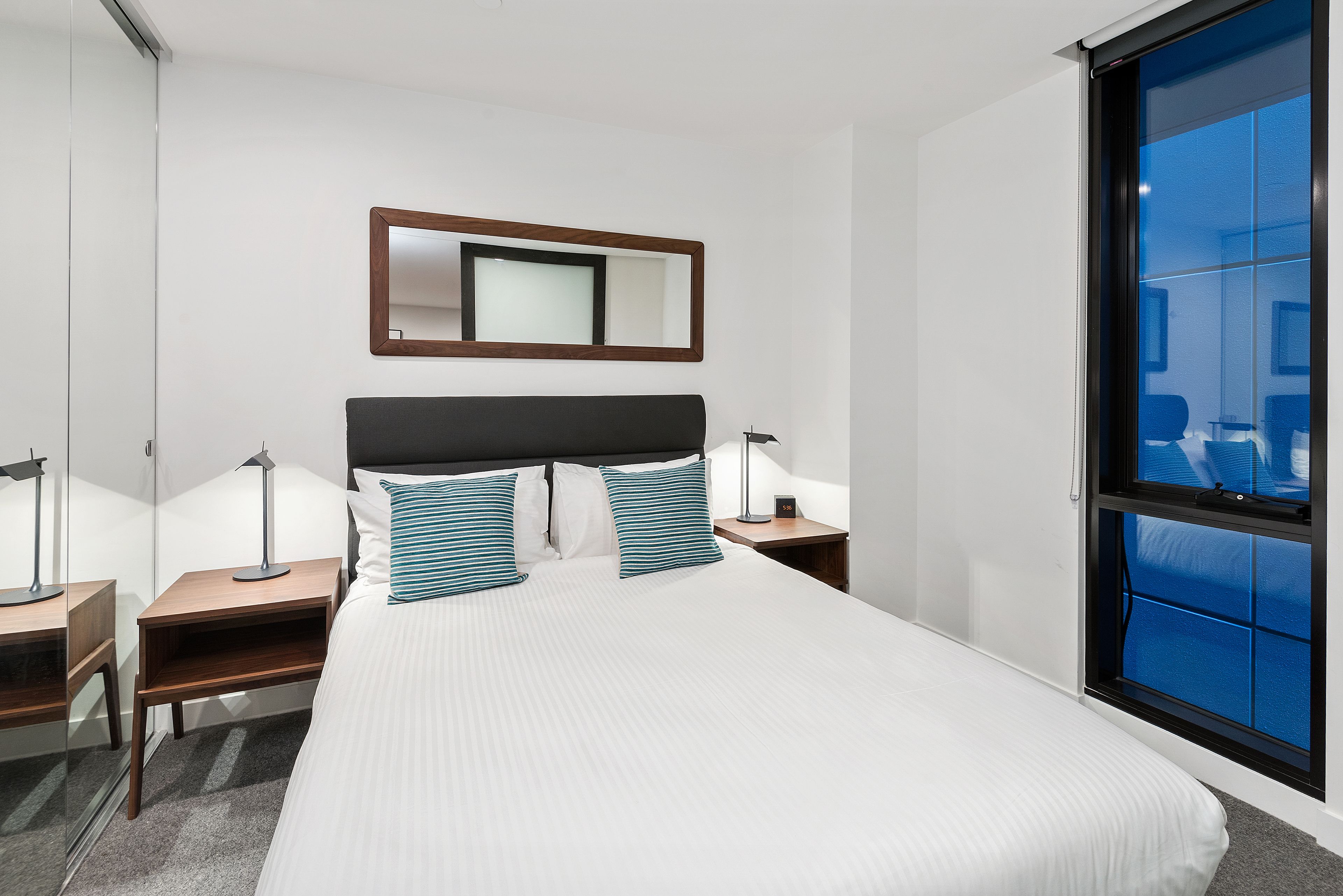 A modern hotel room with a large bed, bedside tables, a mirror, and a window with a view.