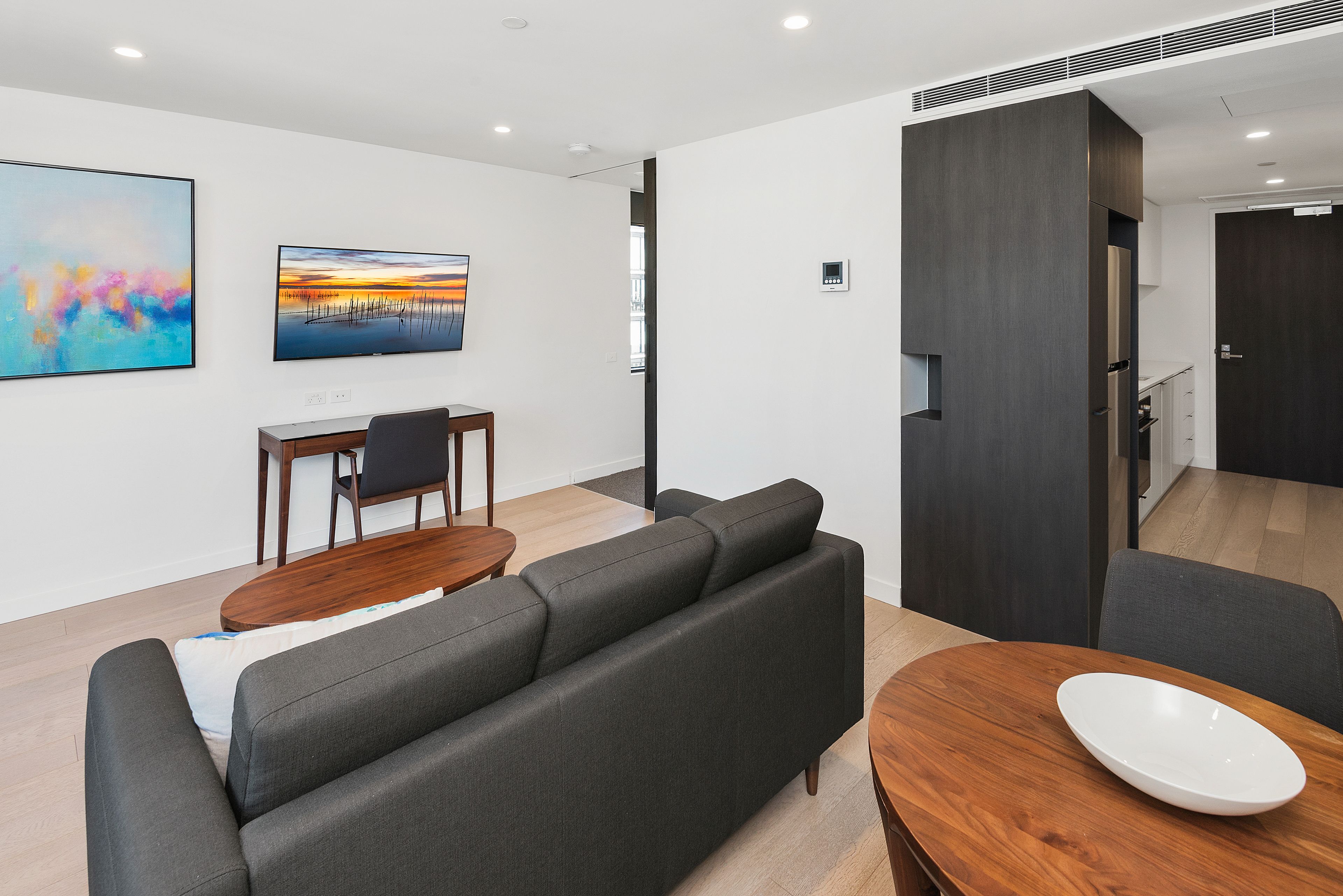 Een moderne woonkamer met een grijze bank, een houten salontafel en een eettafel met stoelen. Twee schilderijen hangen aan de muur en er is een donkere scheidingswand.