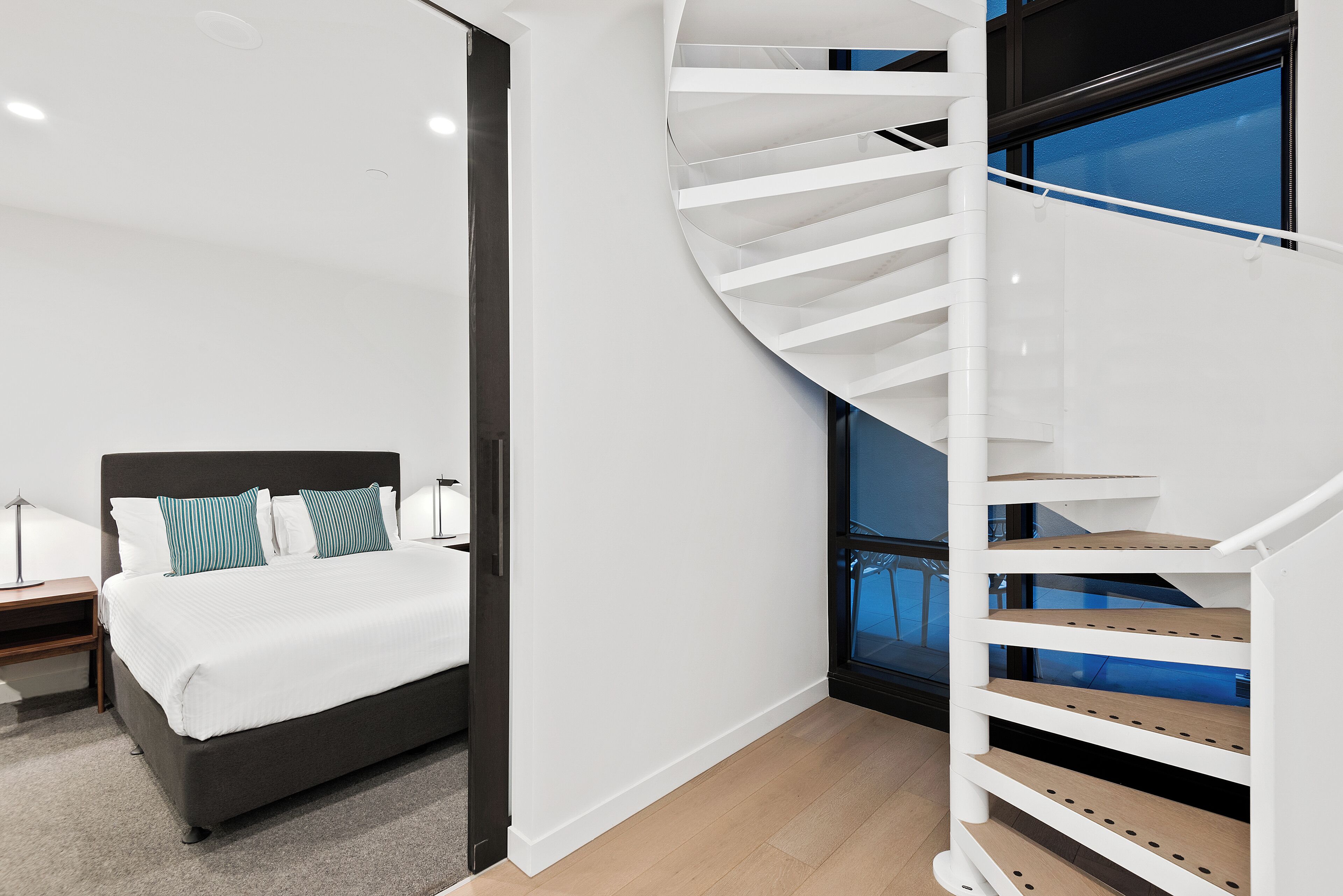 Une chambre à coucher moderne avec un escalier à volutes, un lit recouvert de linge de lit blanc et une table de chevet avec une lampe.