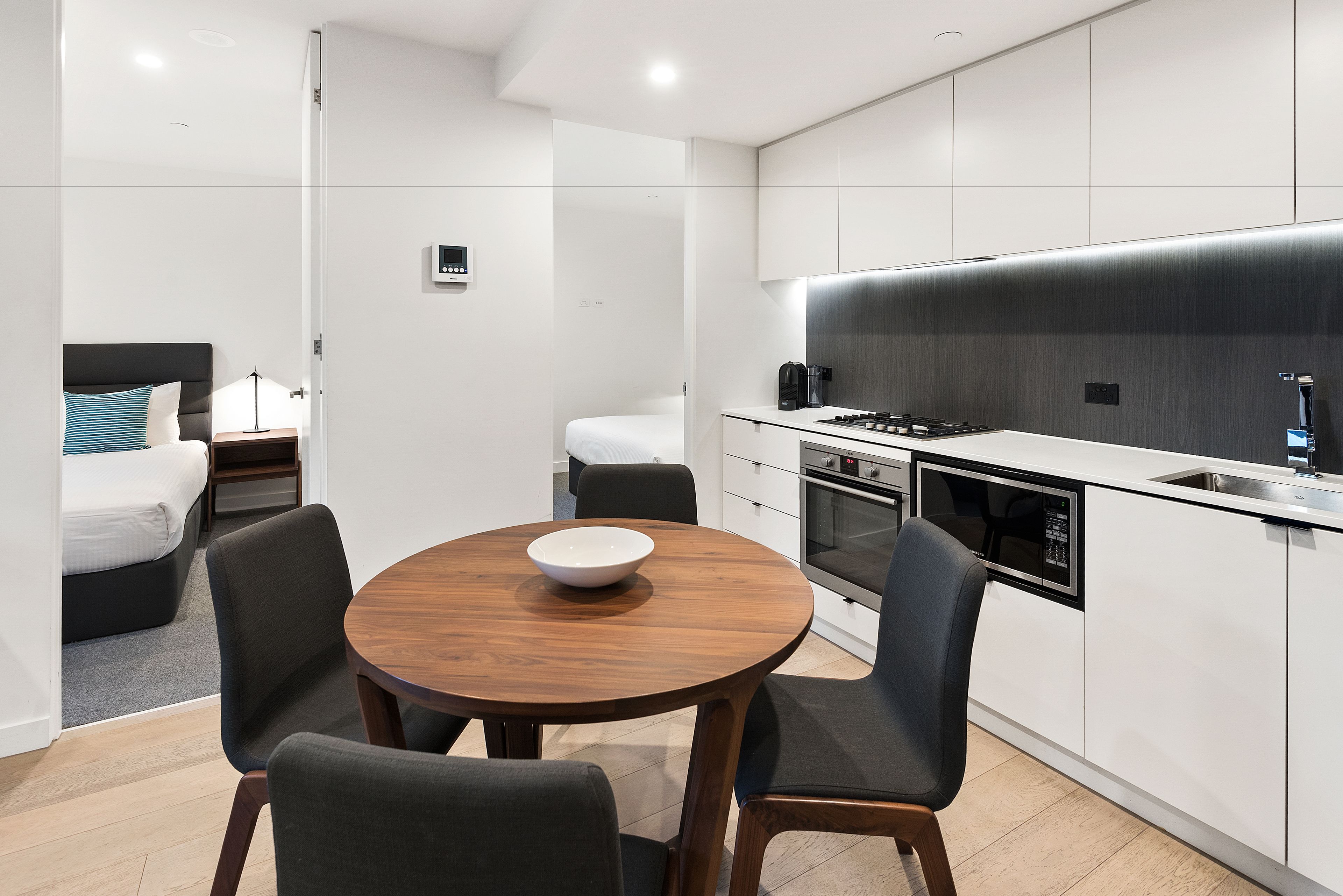 A modern kitchen with white cabinets, a black countertop, and a built-in oven. There is a dining area with a wooden table and four chairs.