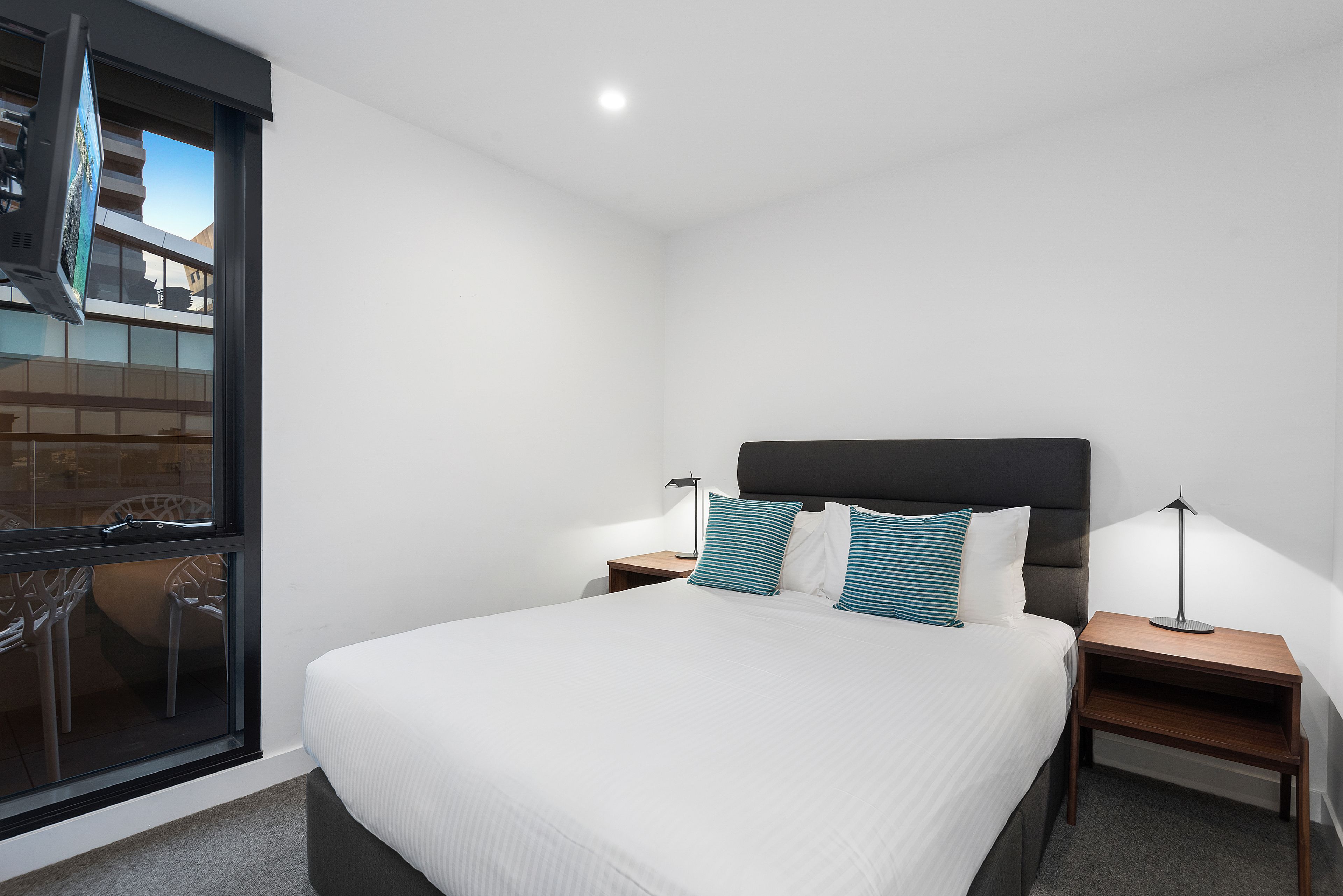 Una habitación de hotel moderna con una cama grande, dos mesitas de noche con lámparas, un ventanal con vistas al exterior y un pequeño balcón.