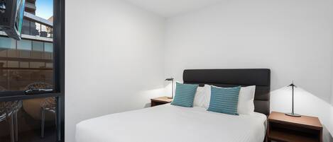 Una habitación de hotel moderna con una cama grande, dos mesitas de noche con lámparas, un ventanal con vistas al exterior y un pequeño balcón.