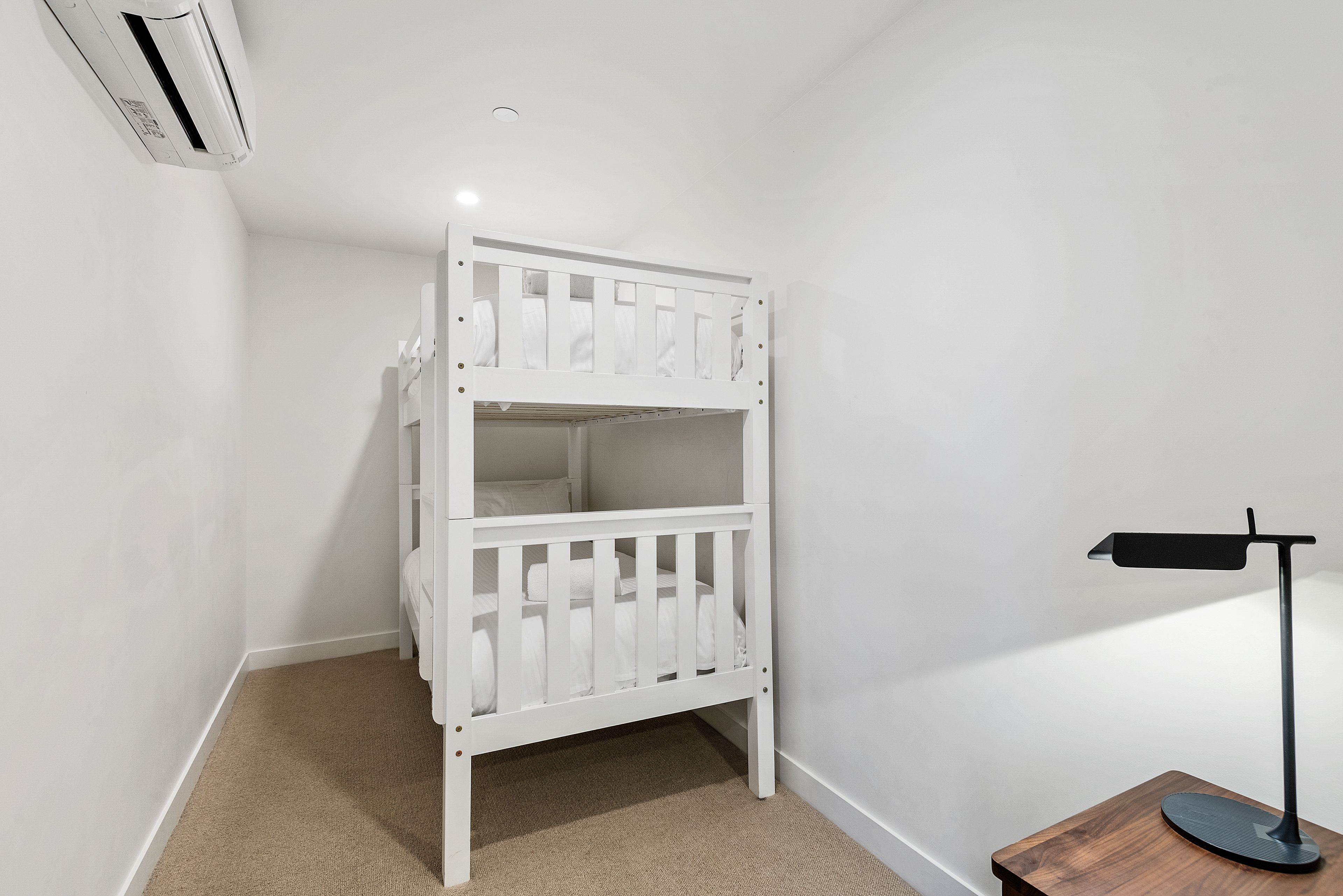 Un lit superposé blanc dans une pièce dont le sol est recouvert d’une moquette, avec une petite table en bois et une lampe, et un appareil de climatisation fixé au mur.