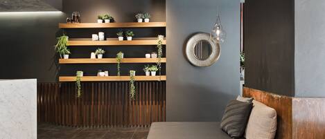 Un área de recepción moderna con un mostrador de madera, estantes con plantas en macetas, un espejo redondo y un banco con cojines.