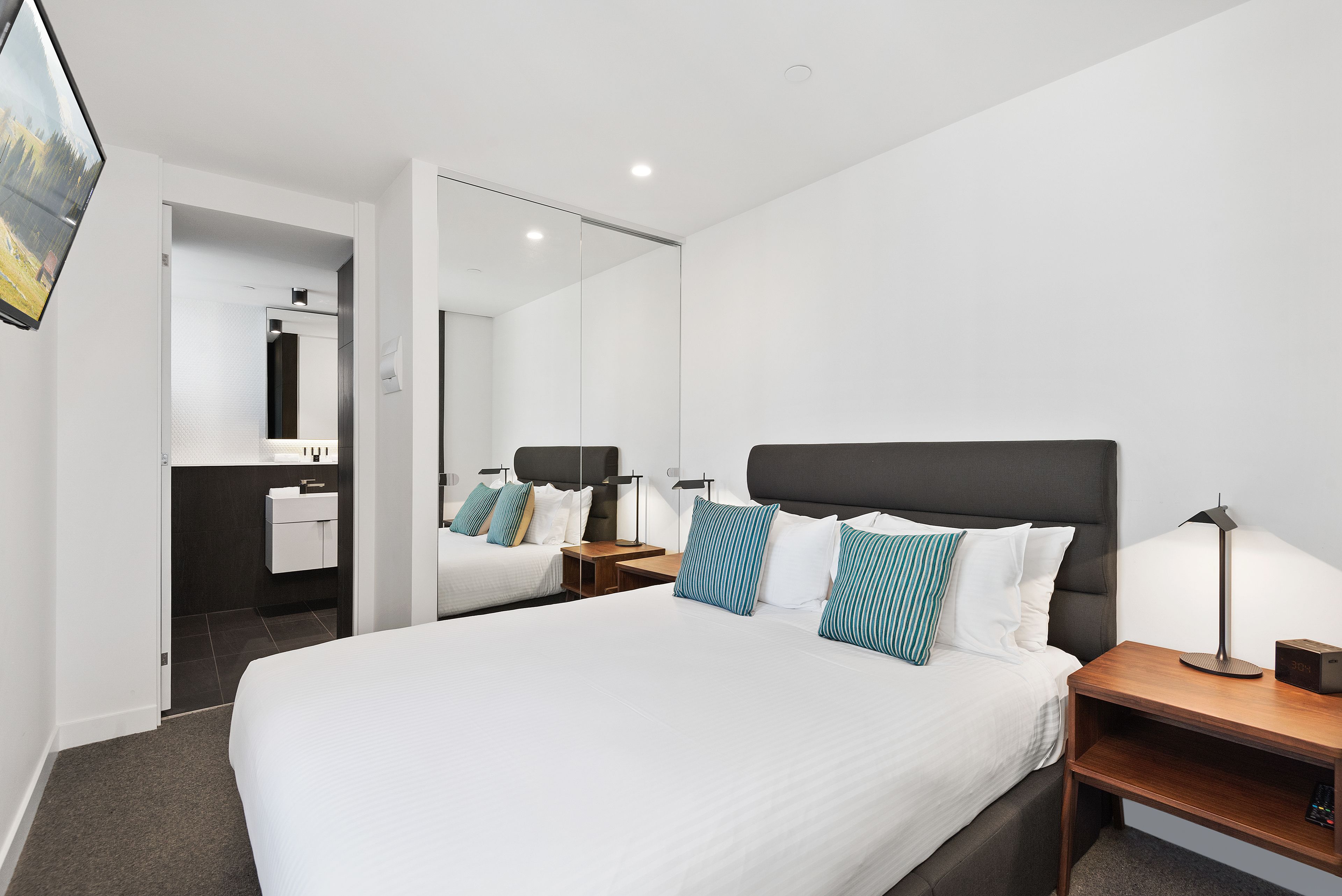 Habitación de hotel moderna con una cama grande, una mesita de noche con lámpara, un espejo y vistas al baño.