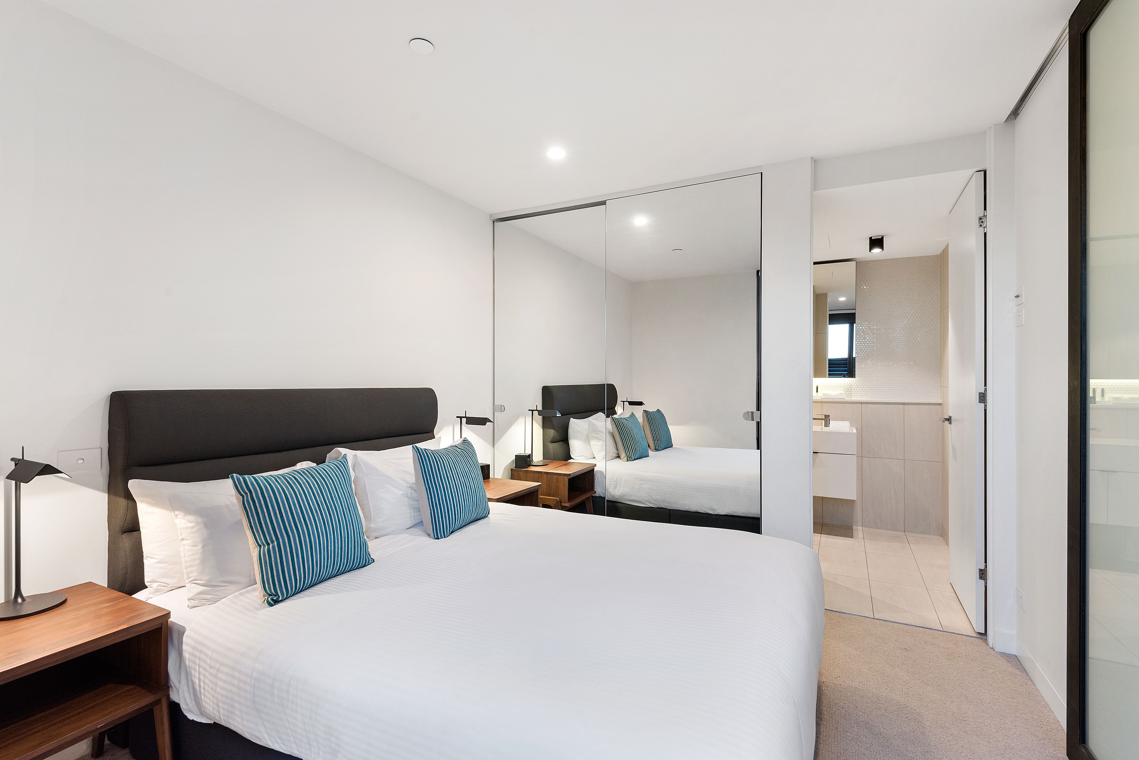 Ein modernes Hotelzimmer mit einem großen Bett, Nachttischen, einem Spiegel und einem durch eine Glastür einsehbaren Badezimmer.