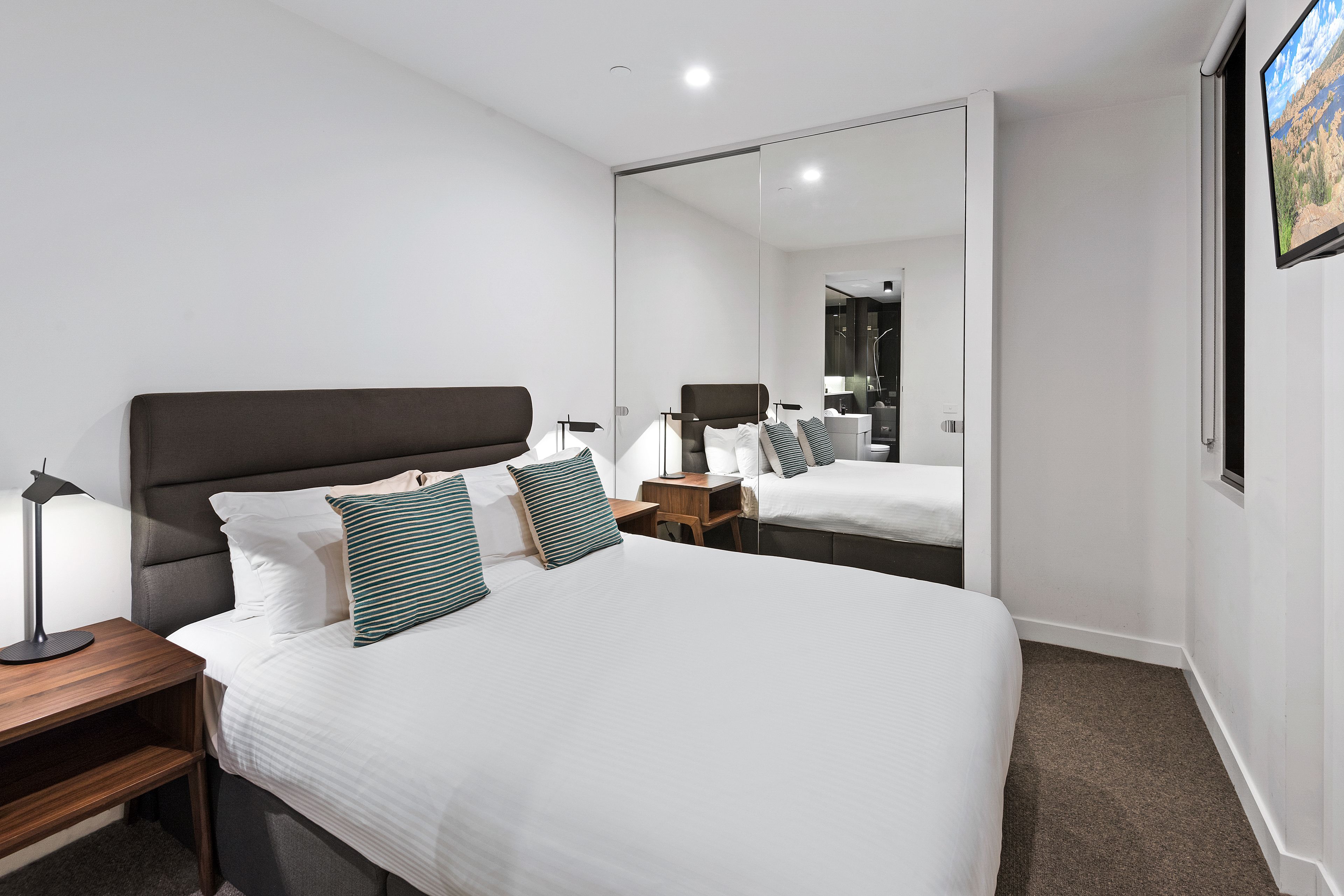 A modern hotel room with a large bed, a bedside table with a lamp, a small wooden nightstand, and a large mirror reflecting the room.